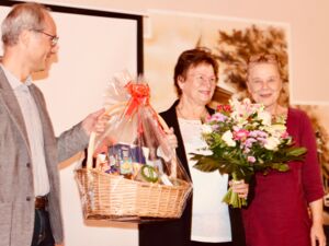 Mann hält einer Frau einen Präsentkorb hin. Die Frau hat in der linken Hand einen großen Blumenstrauß. Daneben steht eine Frau und lächelt in die Kamera.